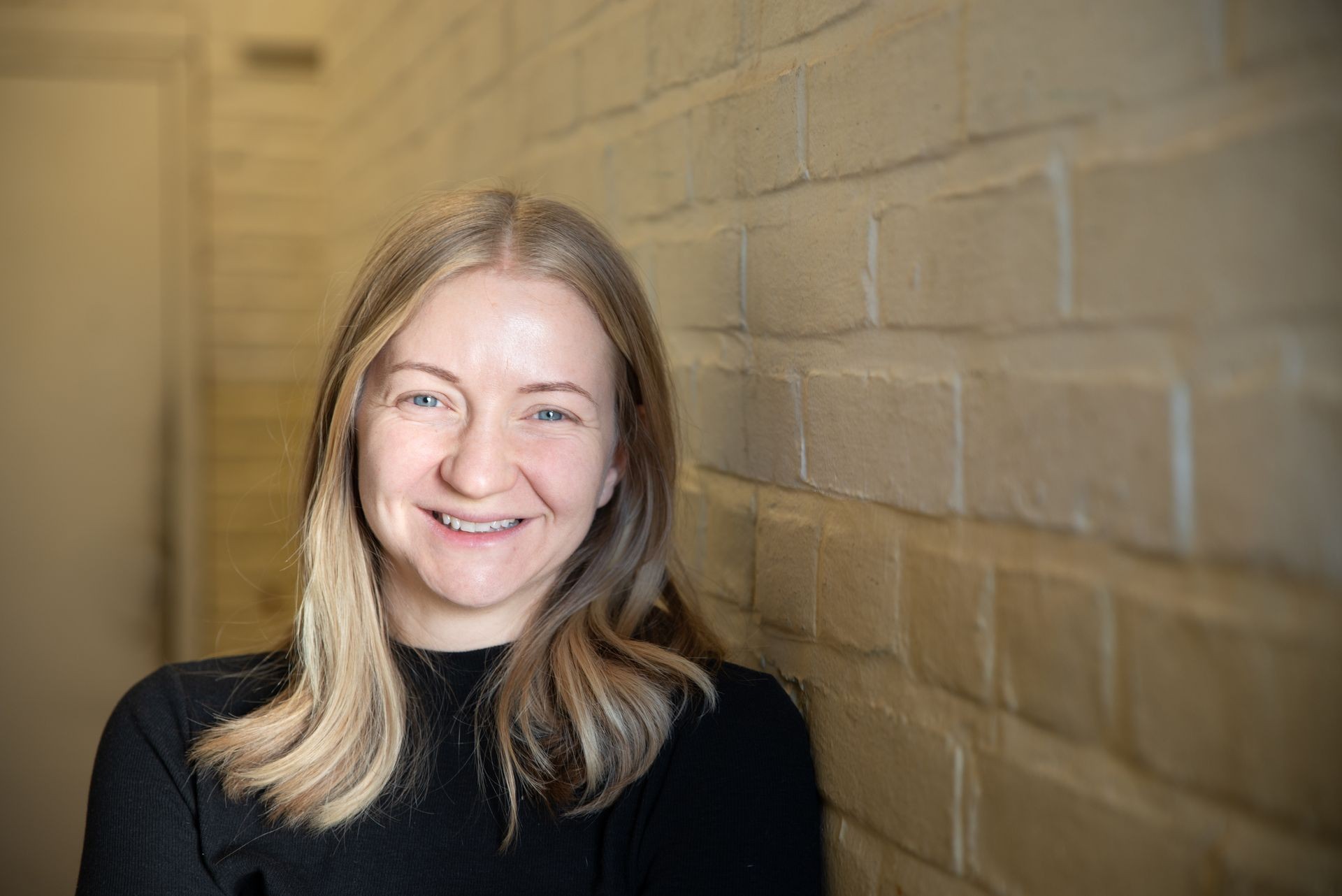 Person with blonde hair wearing a black sweater standing against a beige brick wall.